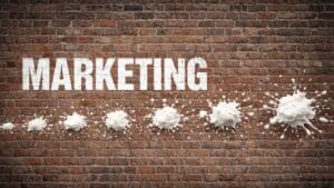 Custard pie style white slop, in a line on a brick wall, as if fired by a machine gun, next to the word 'marketing' in stenciled white letters.